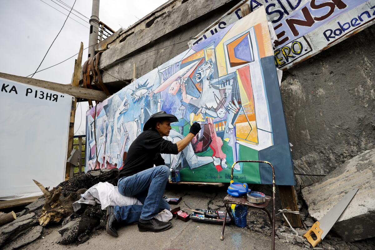 A man in jeans and a black hat, shirt and gloves crouches on one knee next to a painting in front of a damaged bridge 