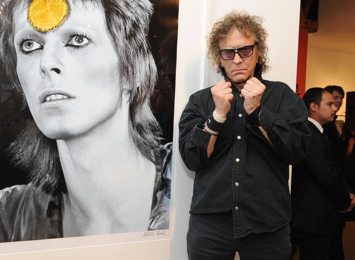 A man in a black button-down shirt stands in front of a photo of David Bowie 