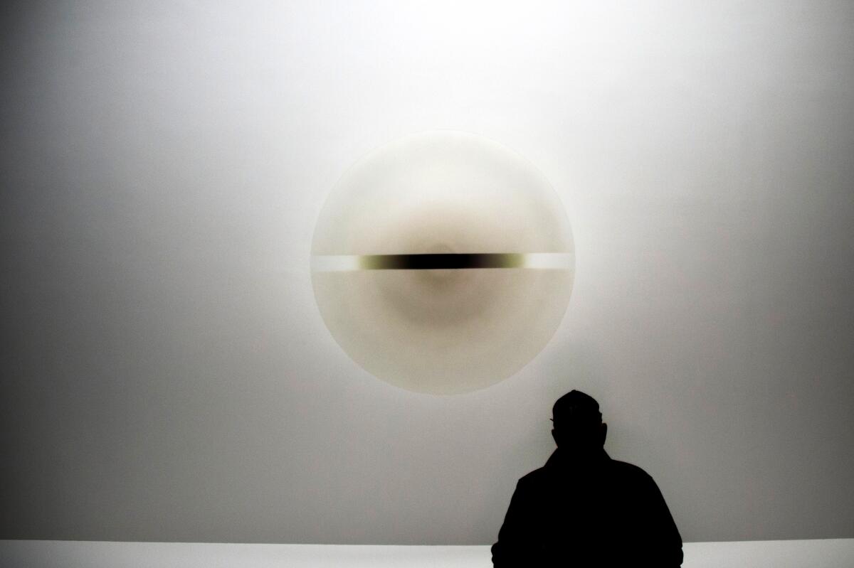 Robert Irwin, in shadow, with his back to the camera, sits in front of his acrylic art.