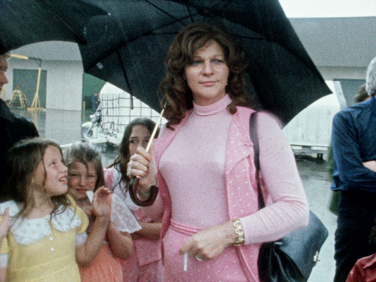 Elizabeth Carmichael in a pink outfit holding an umbrella and a cigarette, accompanied by her family