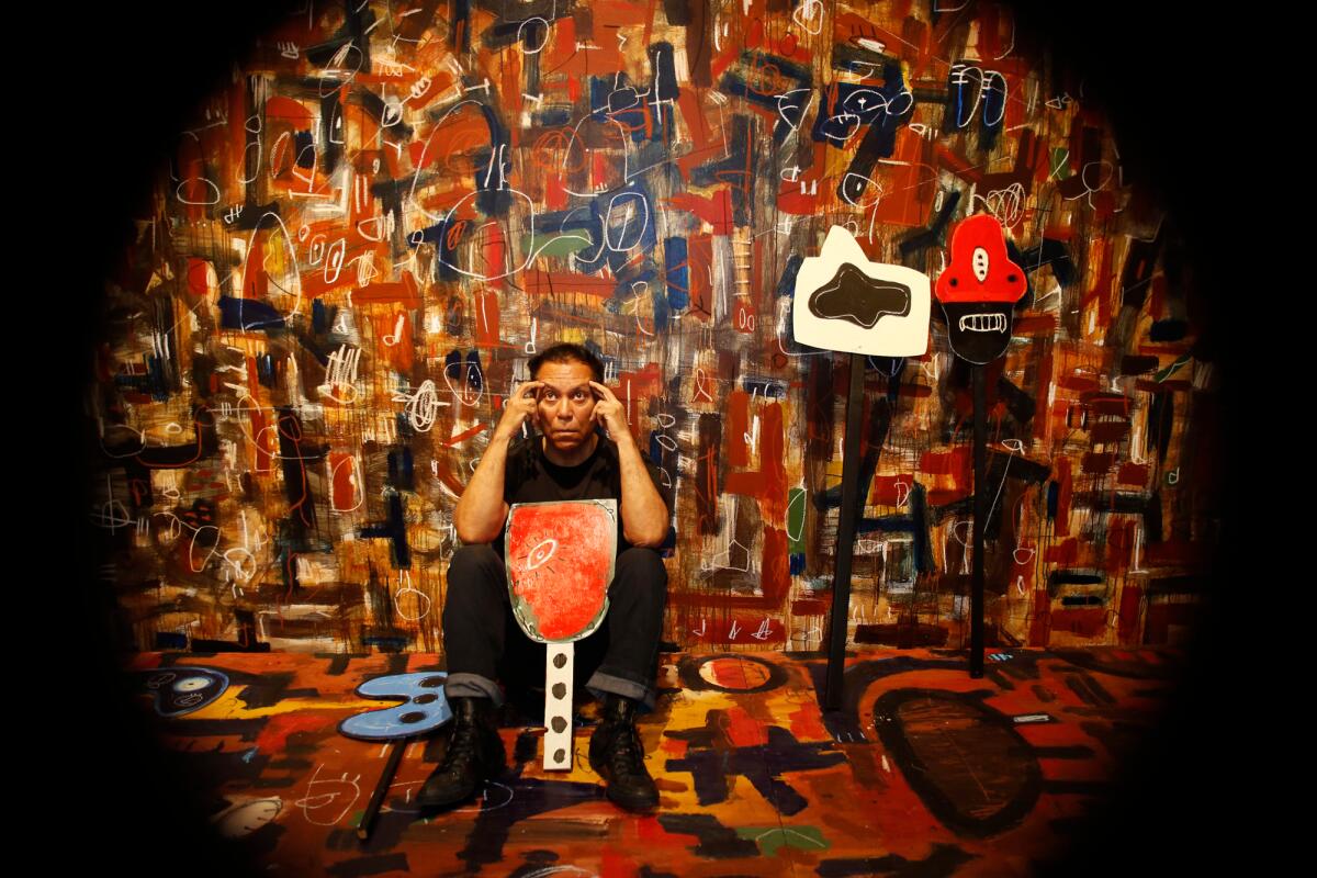 A man sits in a set design that is covered in a painted design featuring black and earth-colored symbols. 