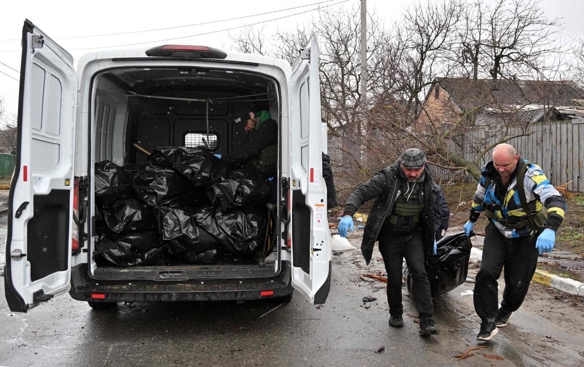 Workers carry a body in a bag to a van carrying other bags. 