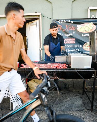 A person wheels a bike past taquero Angel Moreno as he prepares meat for grilling at his taco stand