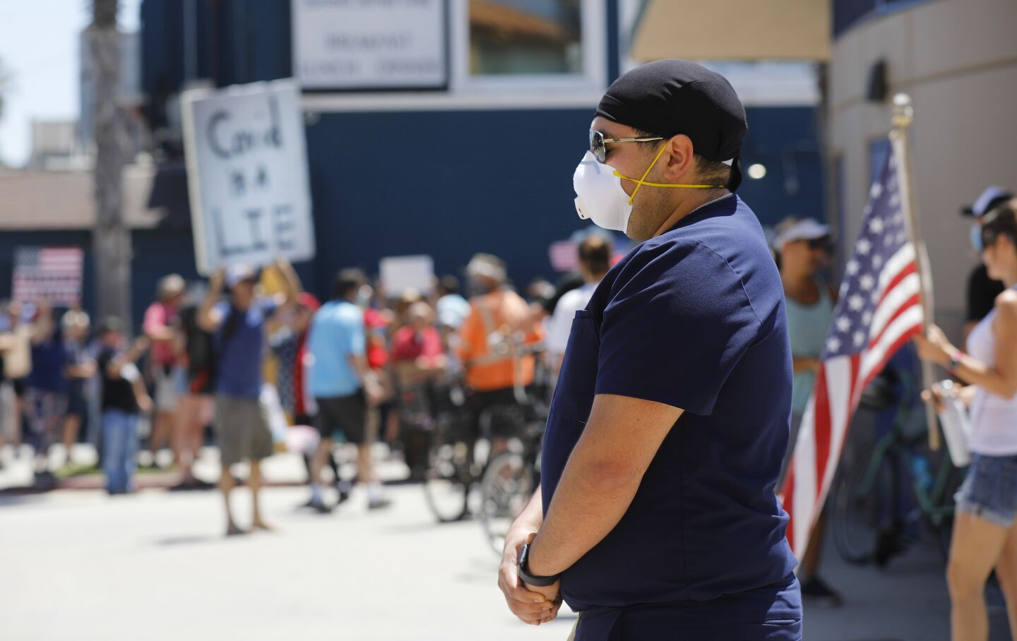 A healthcare worker stands in silence as a protester carries a Covid is a Lie sign during A Day of Liberty rally in Pacific Beach on Sunday, April 26, 2020. The protesters were against the government shutdown due to the coronavirus.