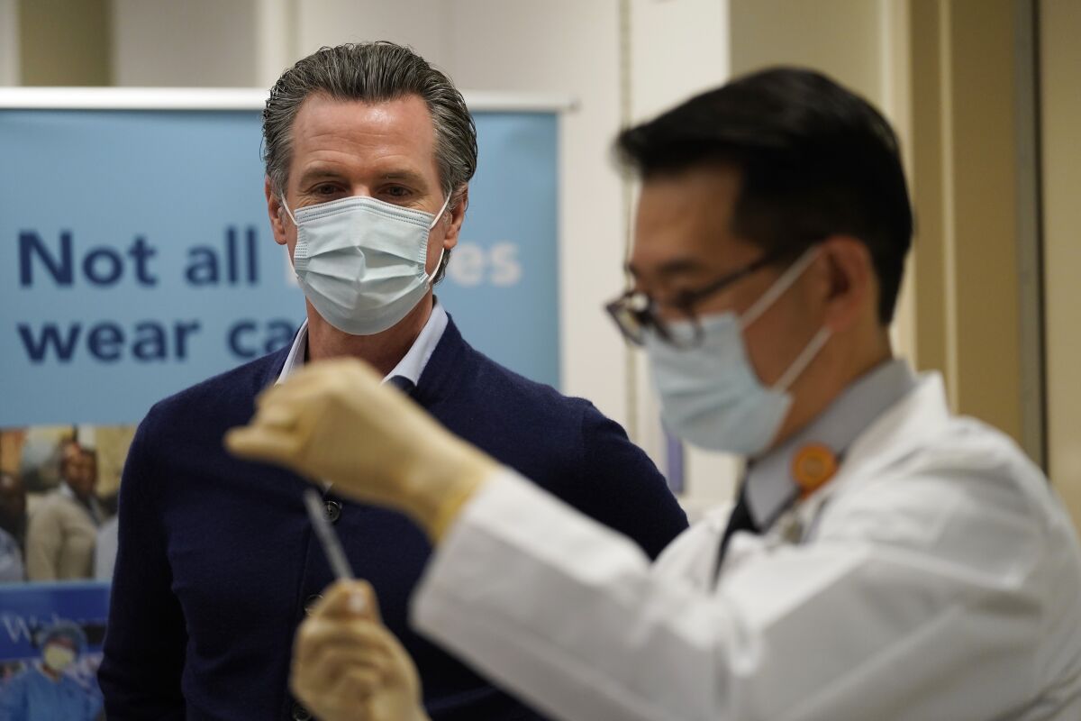 Gavin Newsom in a mask watches a healthcare worker holding up a syringe to prepare a shot
