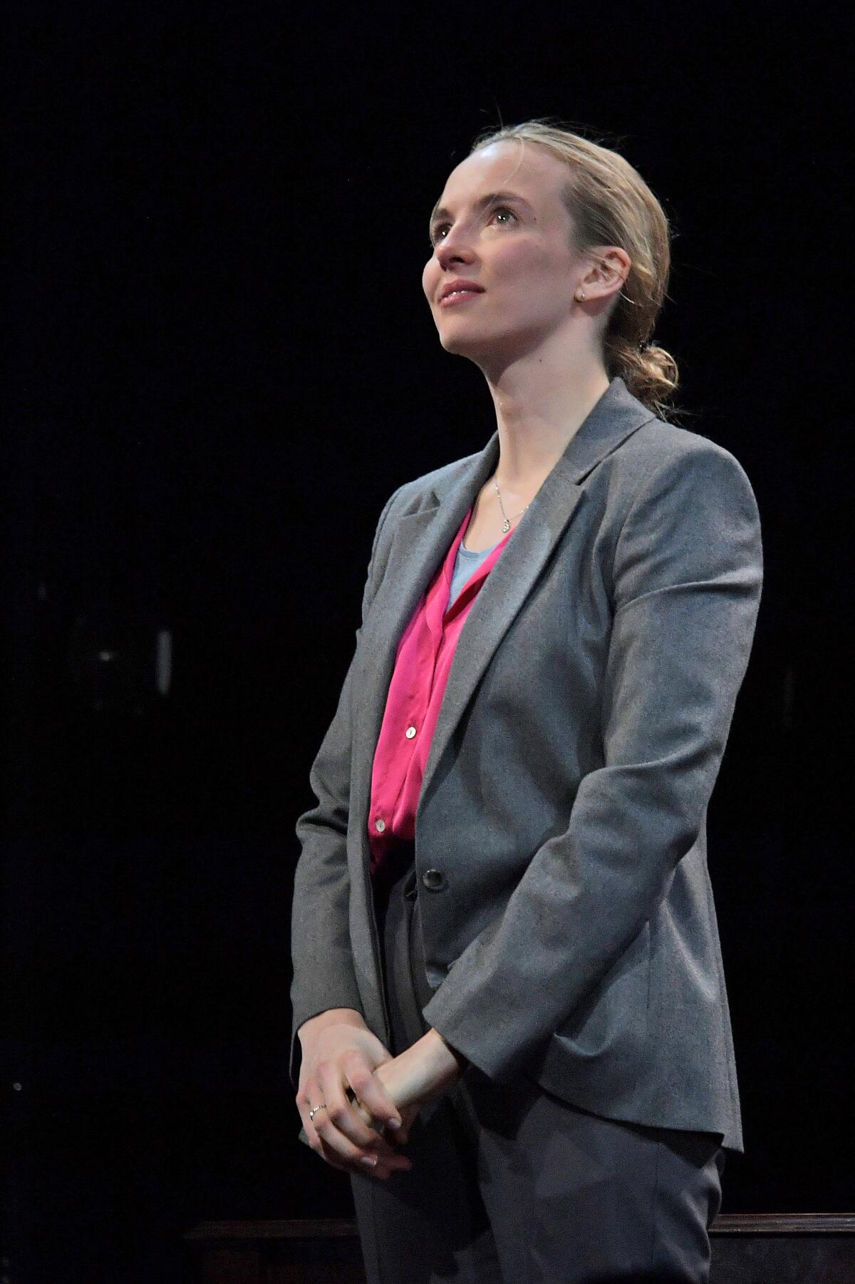 Jodie Comer, in a gray suit and pink shirt, stands with her hands folded on a dark stage.