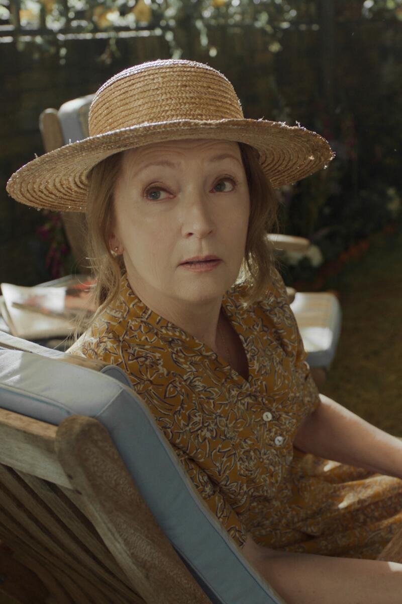 A woman in a sundress and hat sits in an armchair and looks over her shoulder.