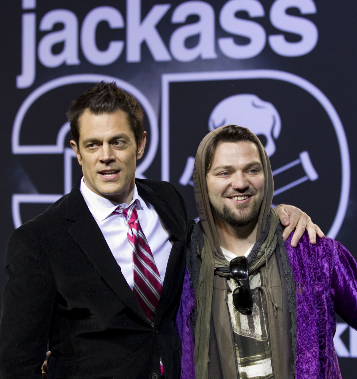 A man in a suit with his arm around a man in a hoodie at a movie premiere.