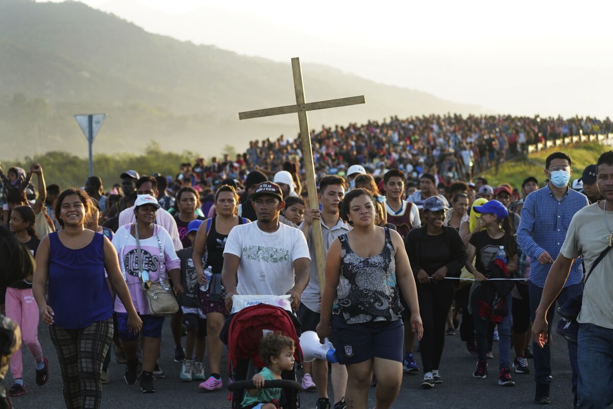 A group of walking migrants, one holding a large cross, stretches back into the distance. 