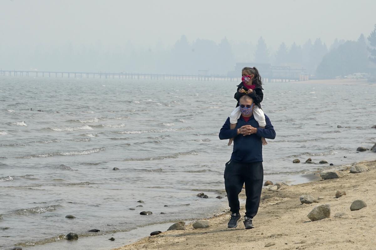 Against the backdrop of a smoky sky, a man carries a girl, both wearing face coverings, on his shoulders at Lake Tahoe.