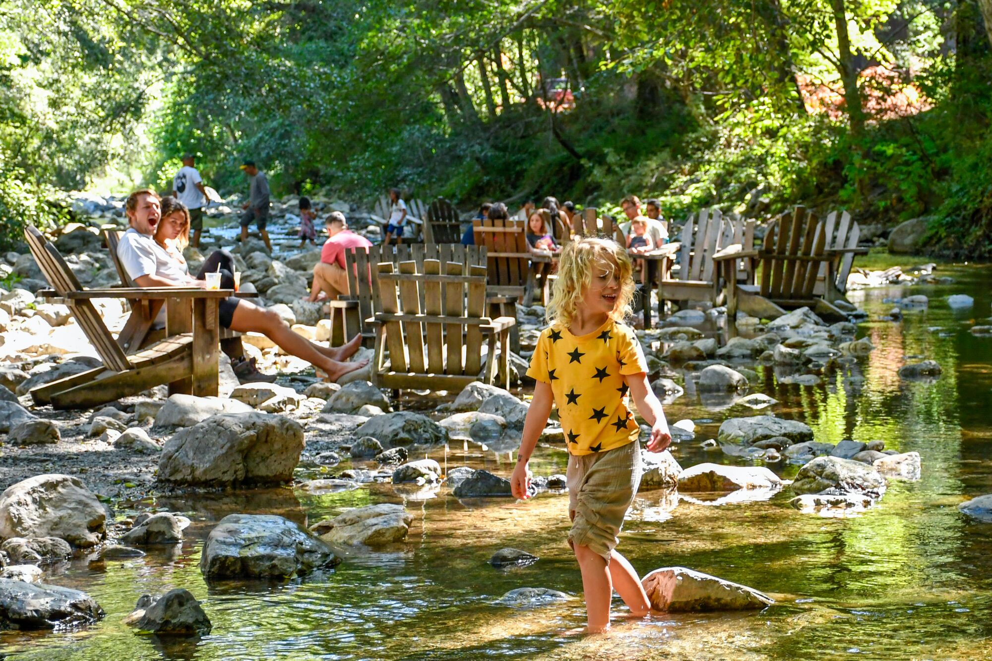 A five-year-old stands ankle-deep in a river, while adults in the background sit in Adirondack chairs, also in the river.