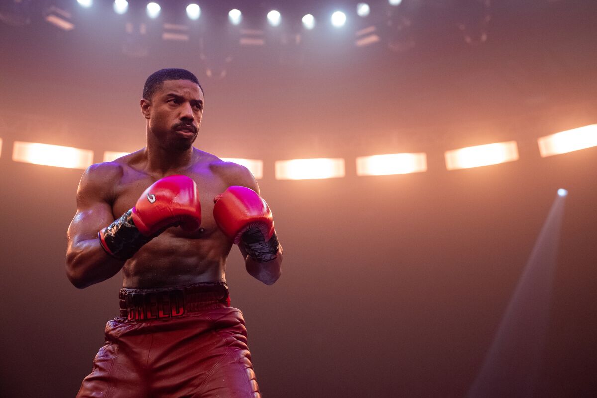 A shirtless man squaring up in red shorts and boxing gloves under stadium lights.