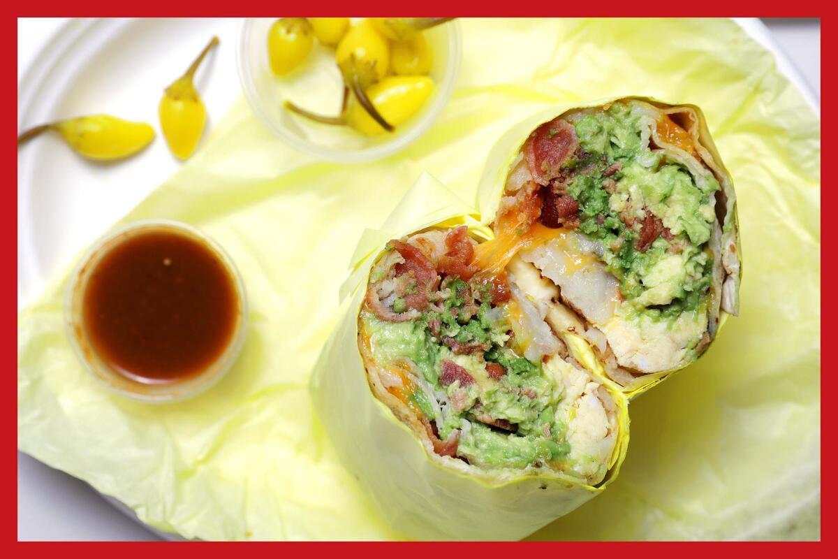 An avocado and bacon breakfast burrito on yellow paper flanked by cups with peppers and sauce