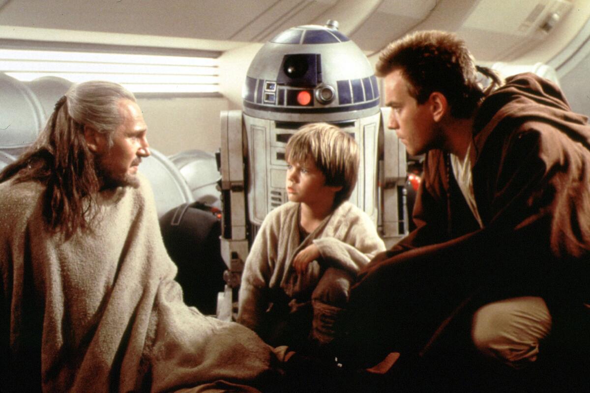 A boy flanked by two men, all dressed in Jedi robes, kneels  near a droid in a scene from a film in the 'Star Wars' franchise