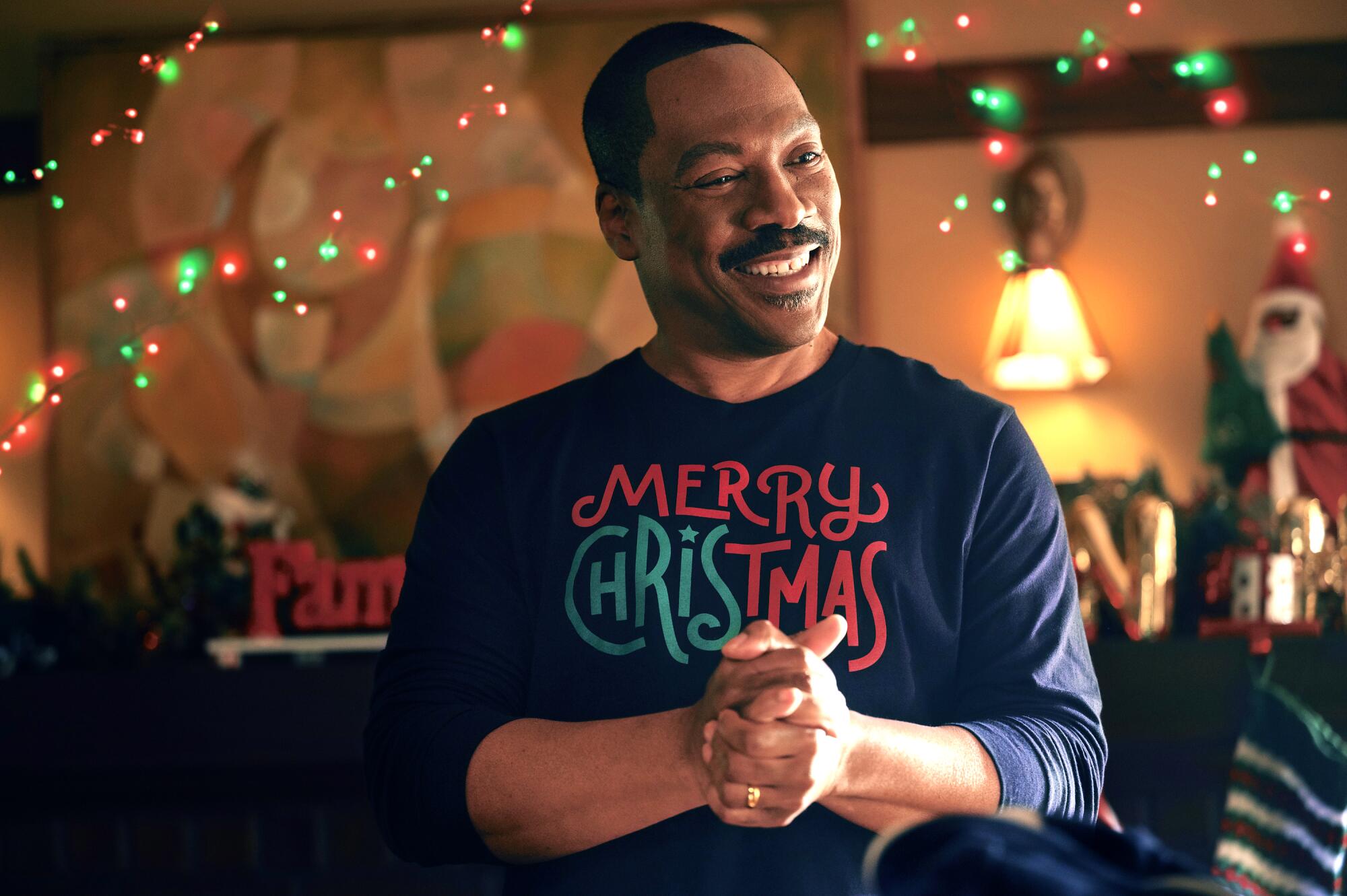 Eddie Murphy as Chris Carver stands with his hands clasped wearing a blue sweater with the words Merry Christmas.