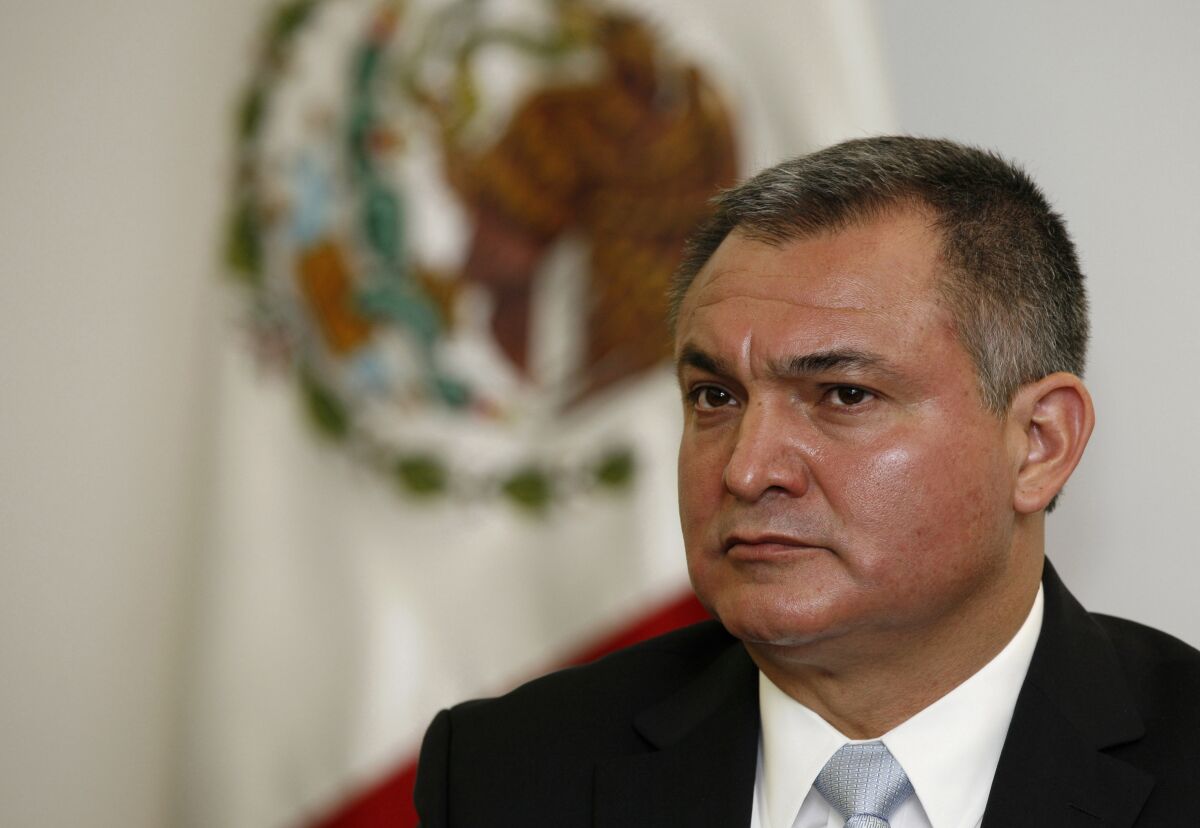 Genaro García Luna is shown sitting in front of a Mexican flag. 