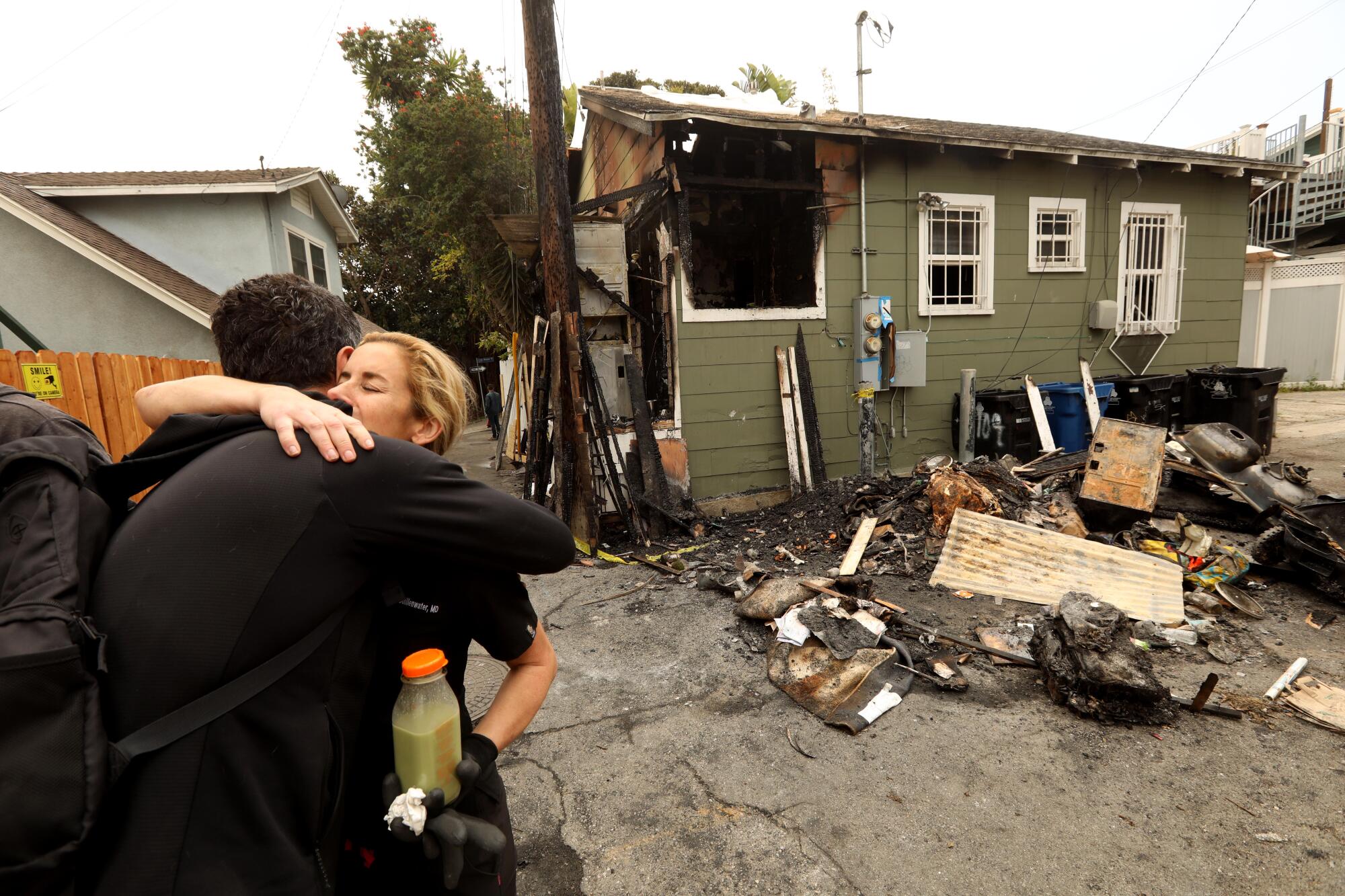 Two people hug outside out of a home with fire damage and debris