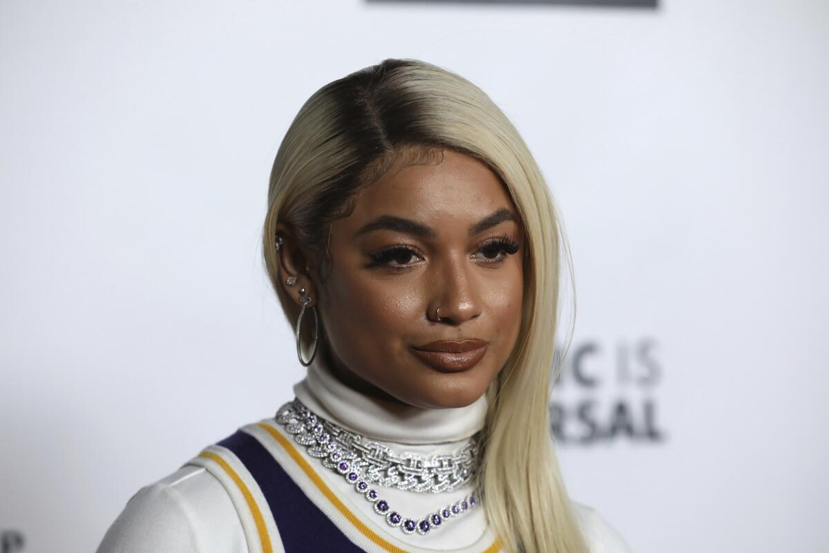 DaniLeigh poses in a white turtleneck and striped shirt with hoop earrings and a purple necklace.