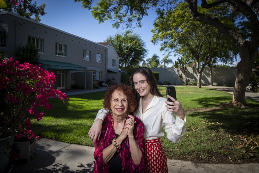 A younger woman drapes her arm around an older woman's shoulders in the yard front of a house.