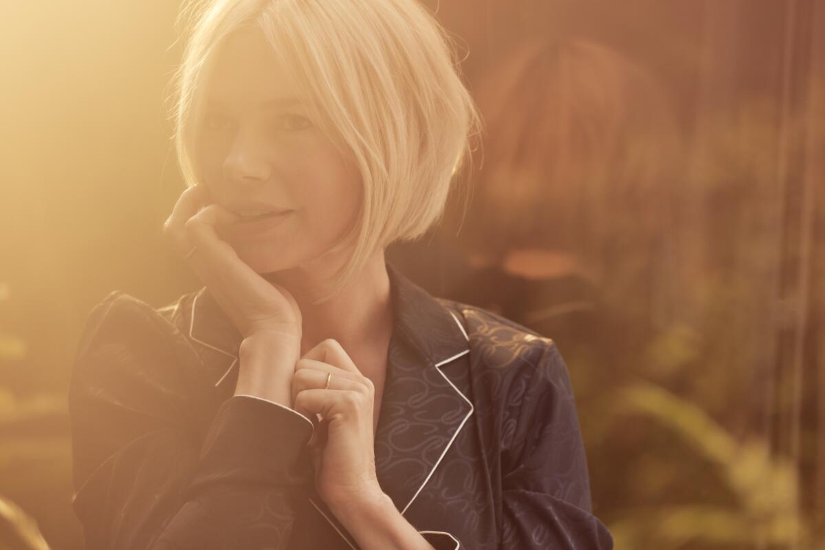 A blond woman stands in an orange sunset's glow, her pinky in her mouth and palm against her chin.