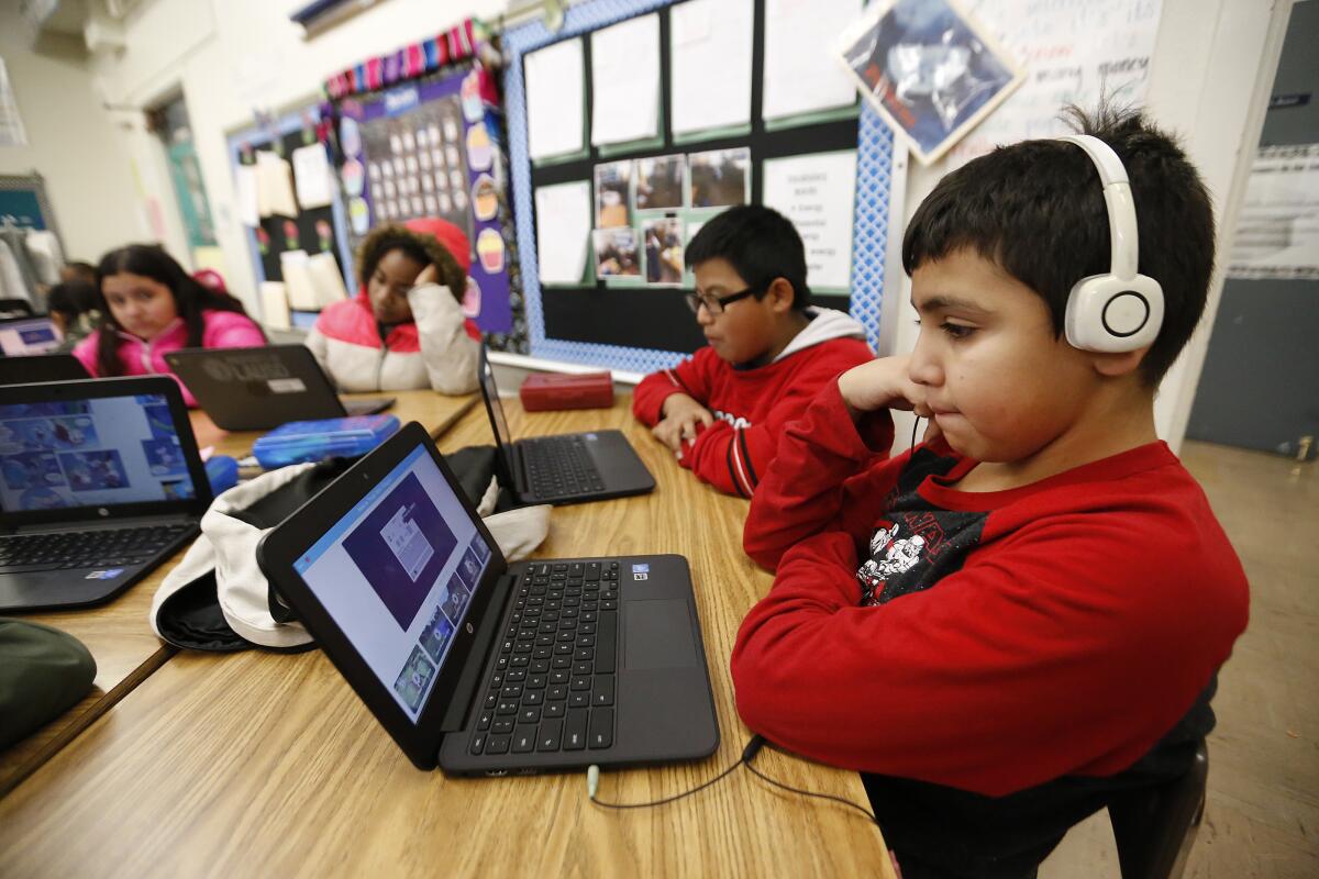 A boy with headphones and three other children look at laptops while sitting at a table in a classroom.