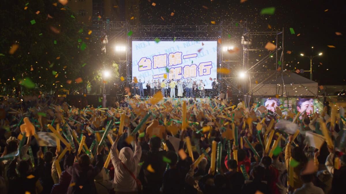 A scene of a political rally with many people, a stage and large video screen behind it in the Netflix show "Wave Makers"