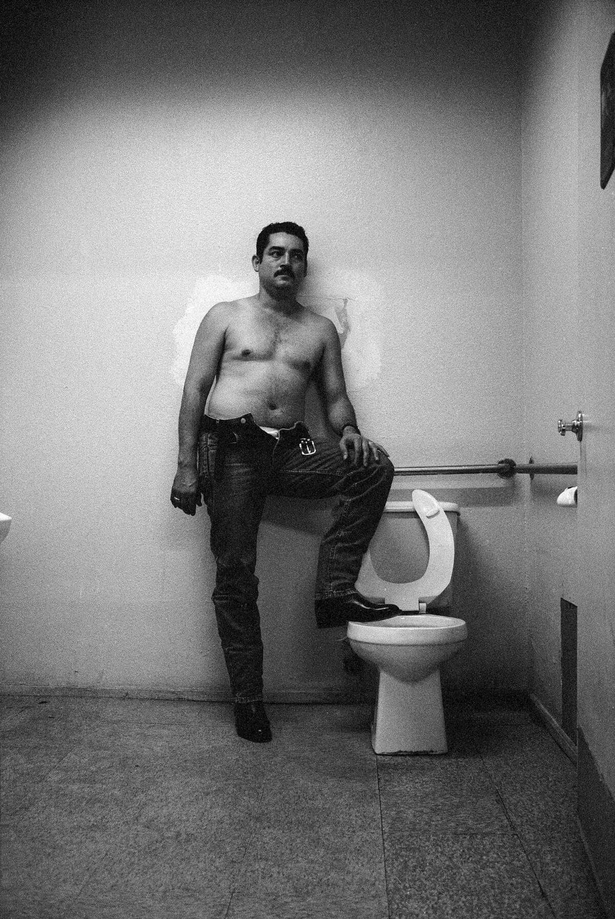A shirtless man stands in a bathroom, his foot jauntily perched on the edge of a toilet