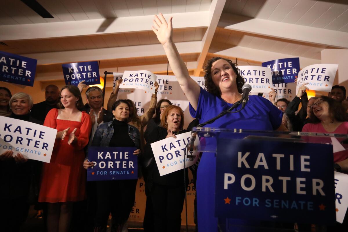 U.S. Representative Katie Porter stands waving from a podium, with supporters behind her holding Katie Porter signs