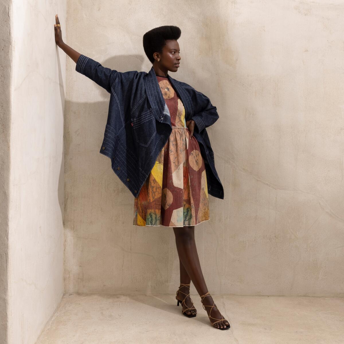 A woman leans against a wall in a denim jacket and patterned dress.