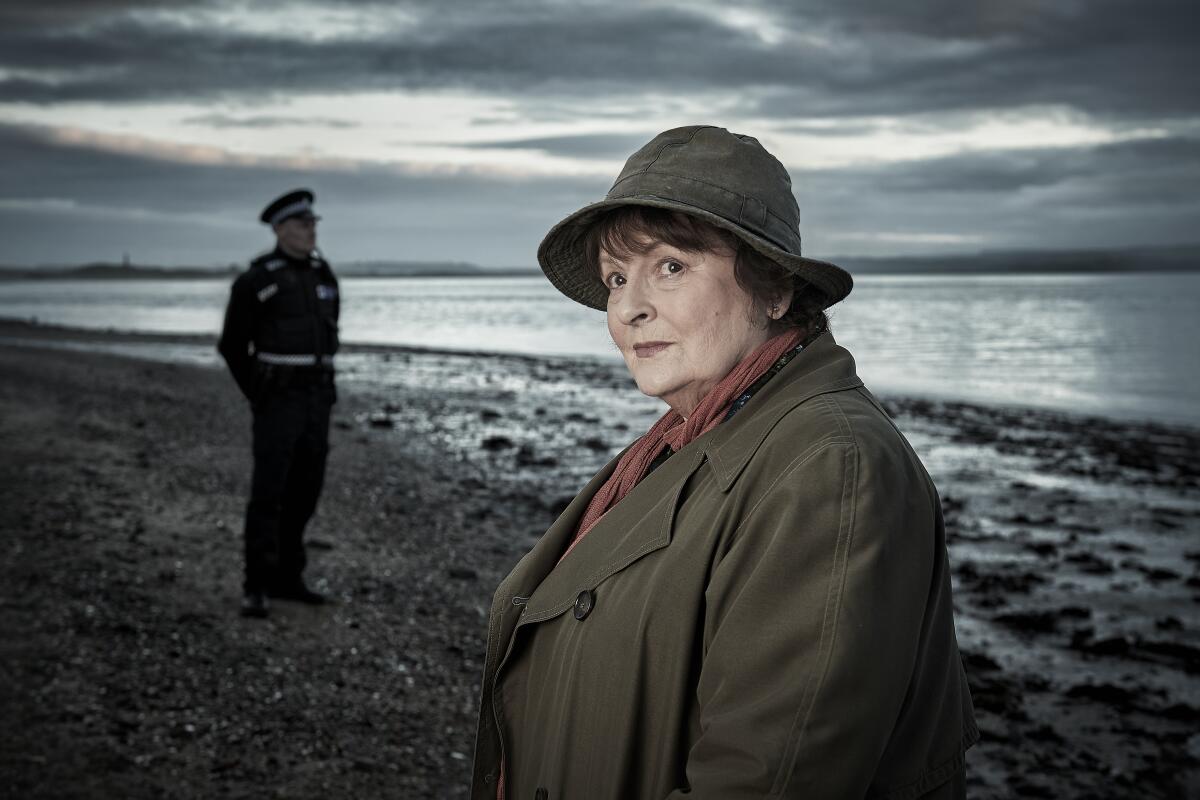 A woman detective stands on a beach with a male police officer.