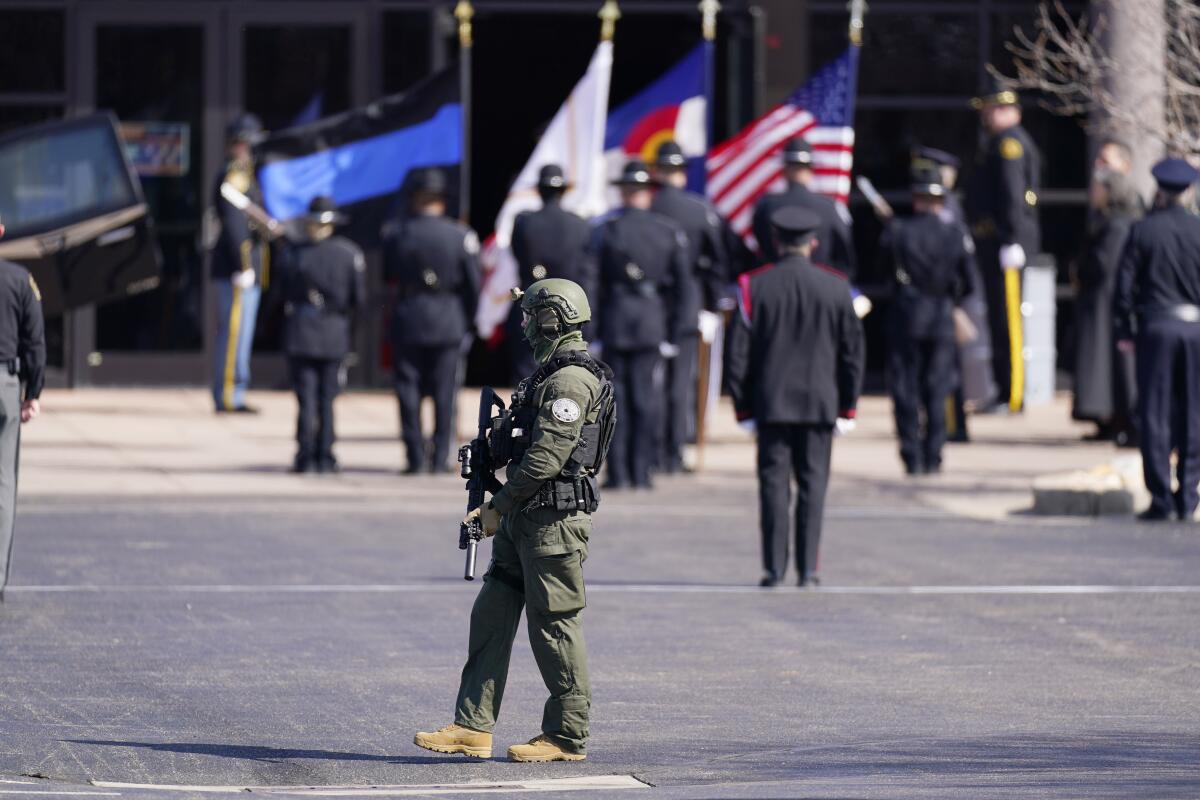 An officer carrying a gun walks in a parking lot; in the background, officers stand at attention.