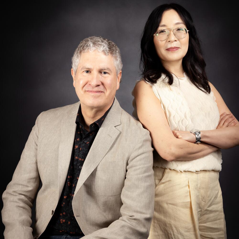 A man in a tan blazer sits and poses for a photo next to a woman, arms crossed, wearing a cream colored top and tan pants.