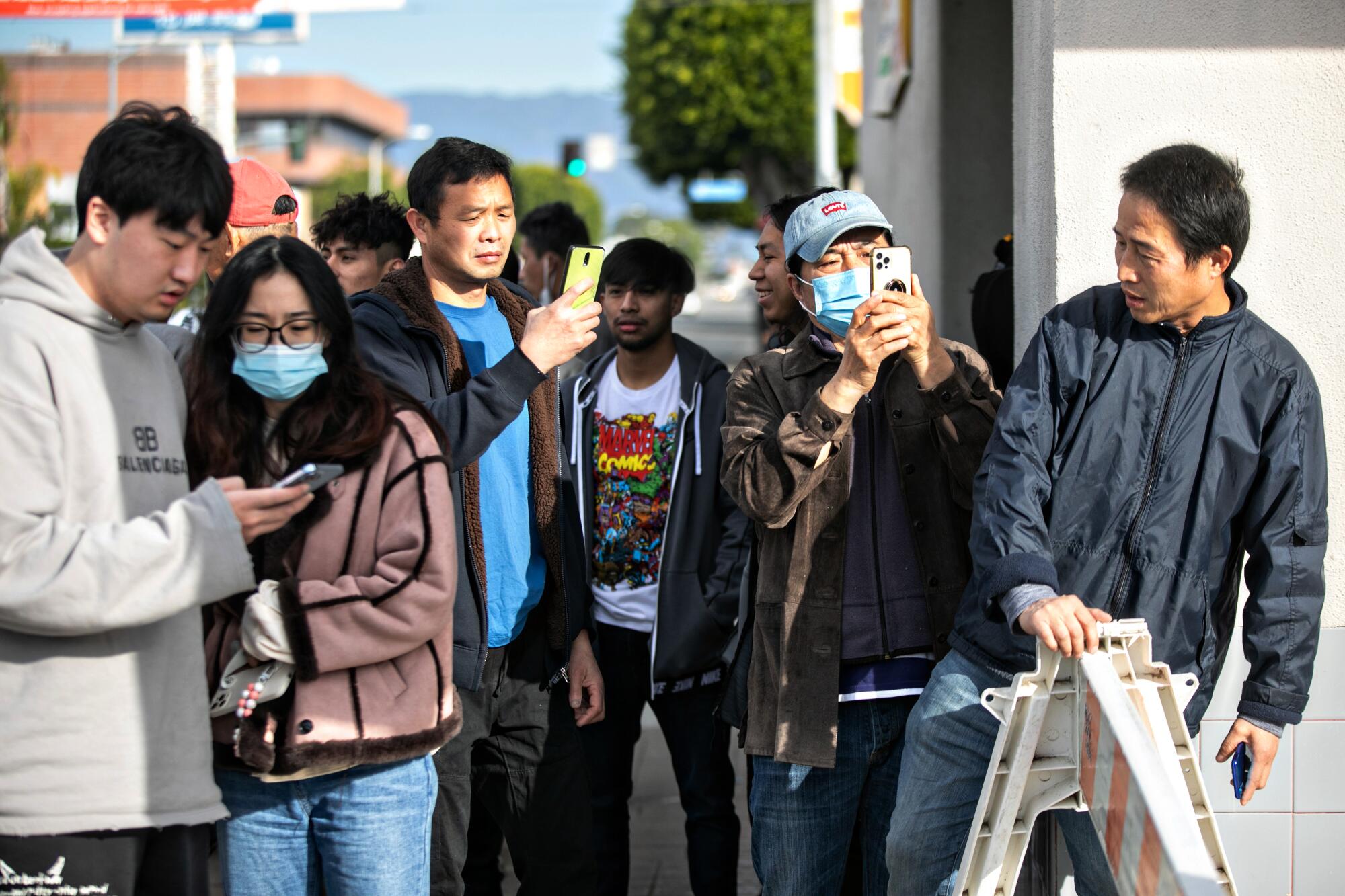 A small group of people stands on a street, some having their phones out as if recording.