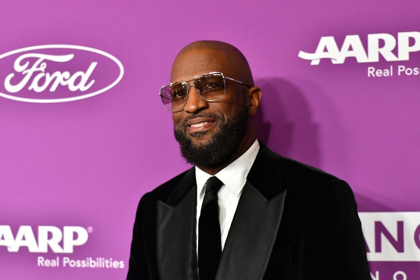 A bald man with a beard smiles while wearing sunglasses and a suit