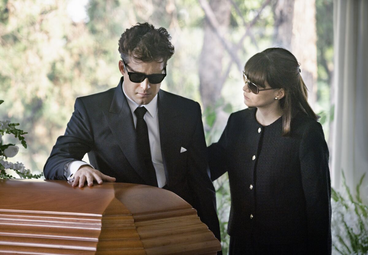 A man in a black suit and sunglasses is comforted by a woman as he places his hand on a casket.