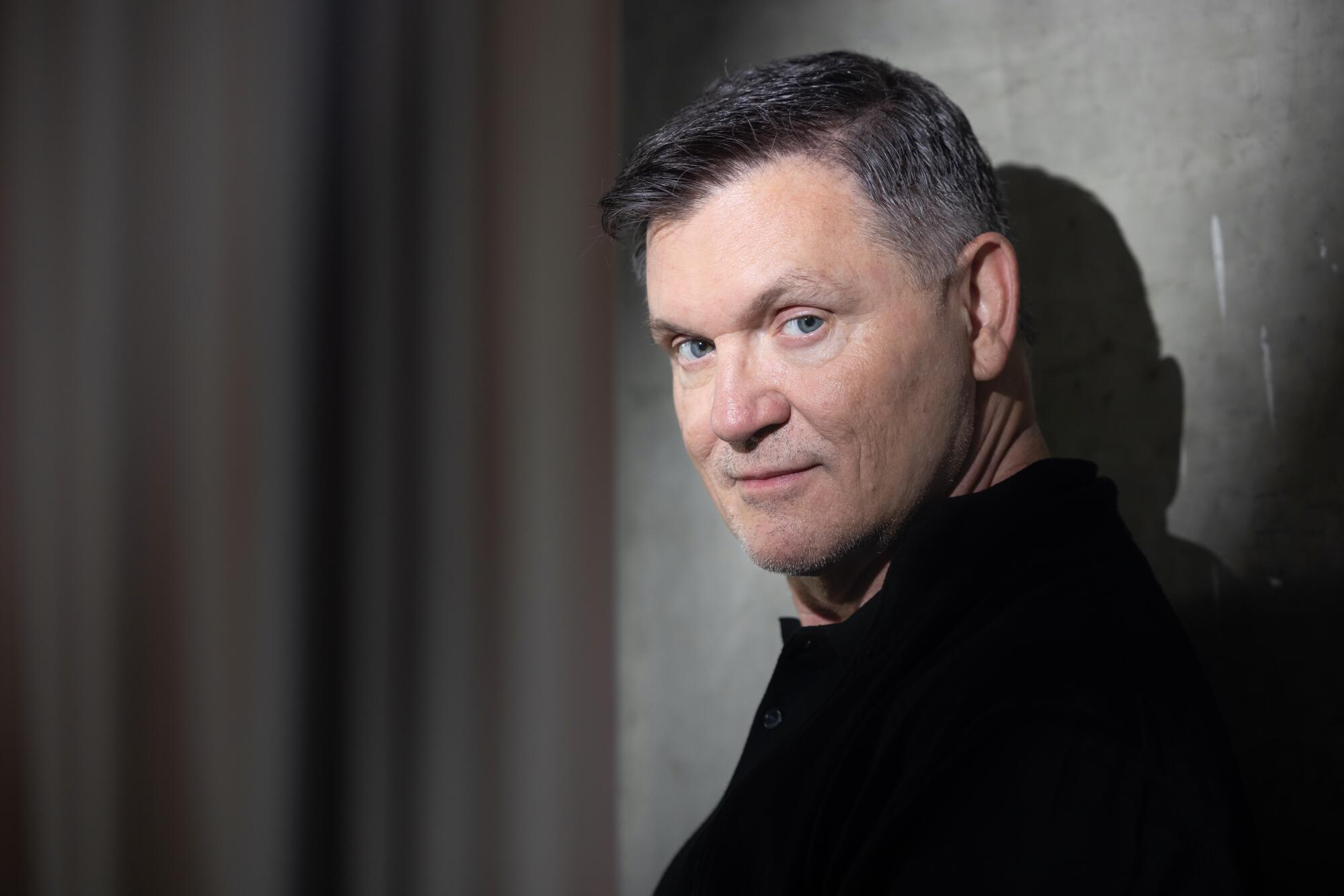 A man with short greying hair in a dark shirt, leaning against a wall.