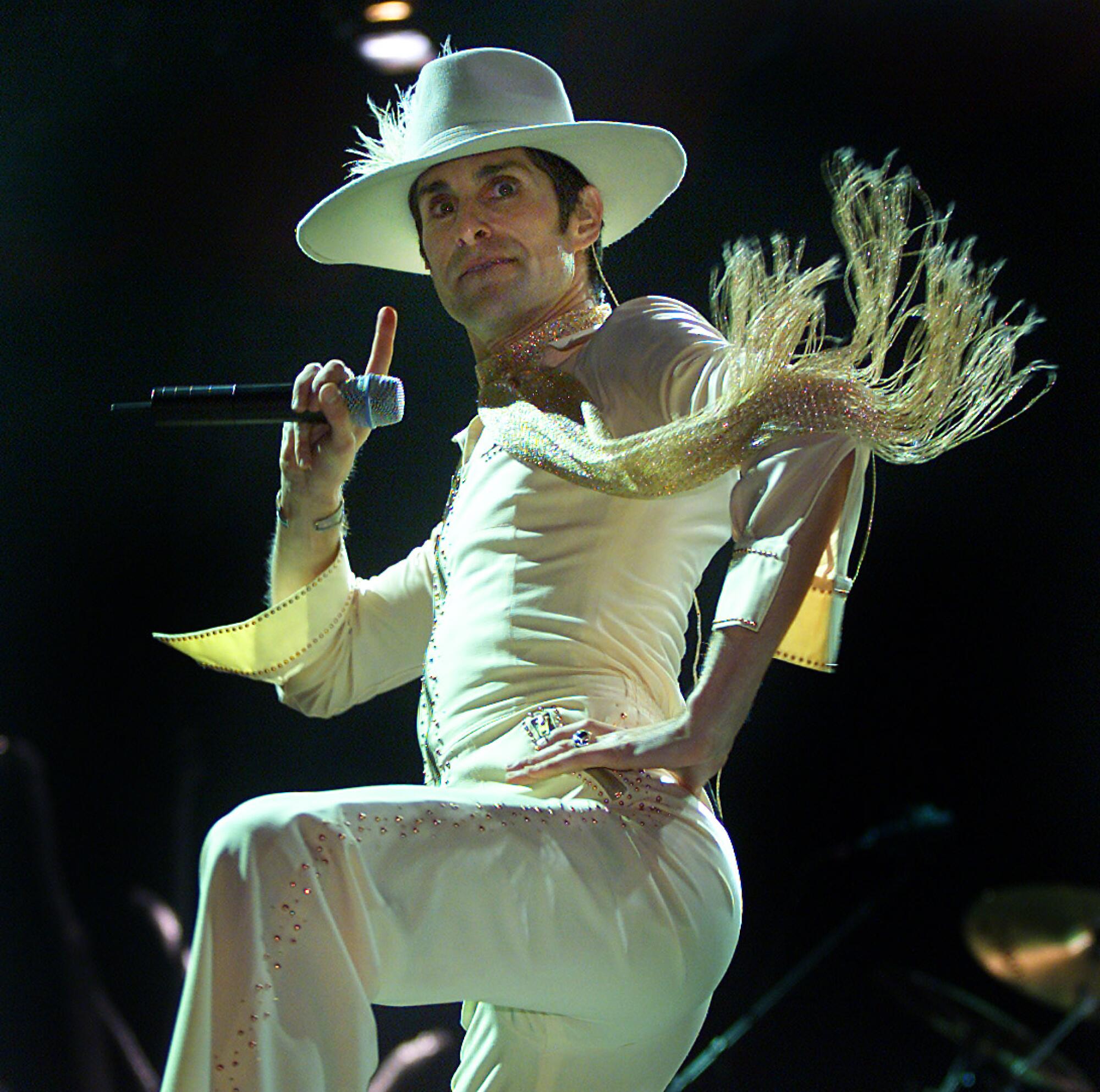 Perry Farrell holds a microphone portion    wearing a achromatic  flowy outfit with fringe and a ample  hat