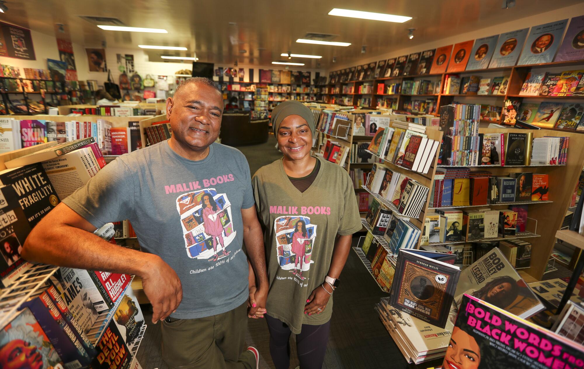A man and a woman posing in "Malik Books" shirts in a bookstore.