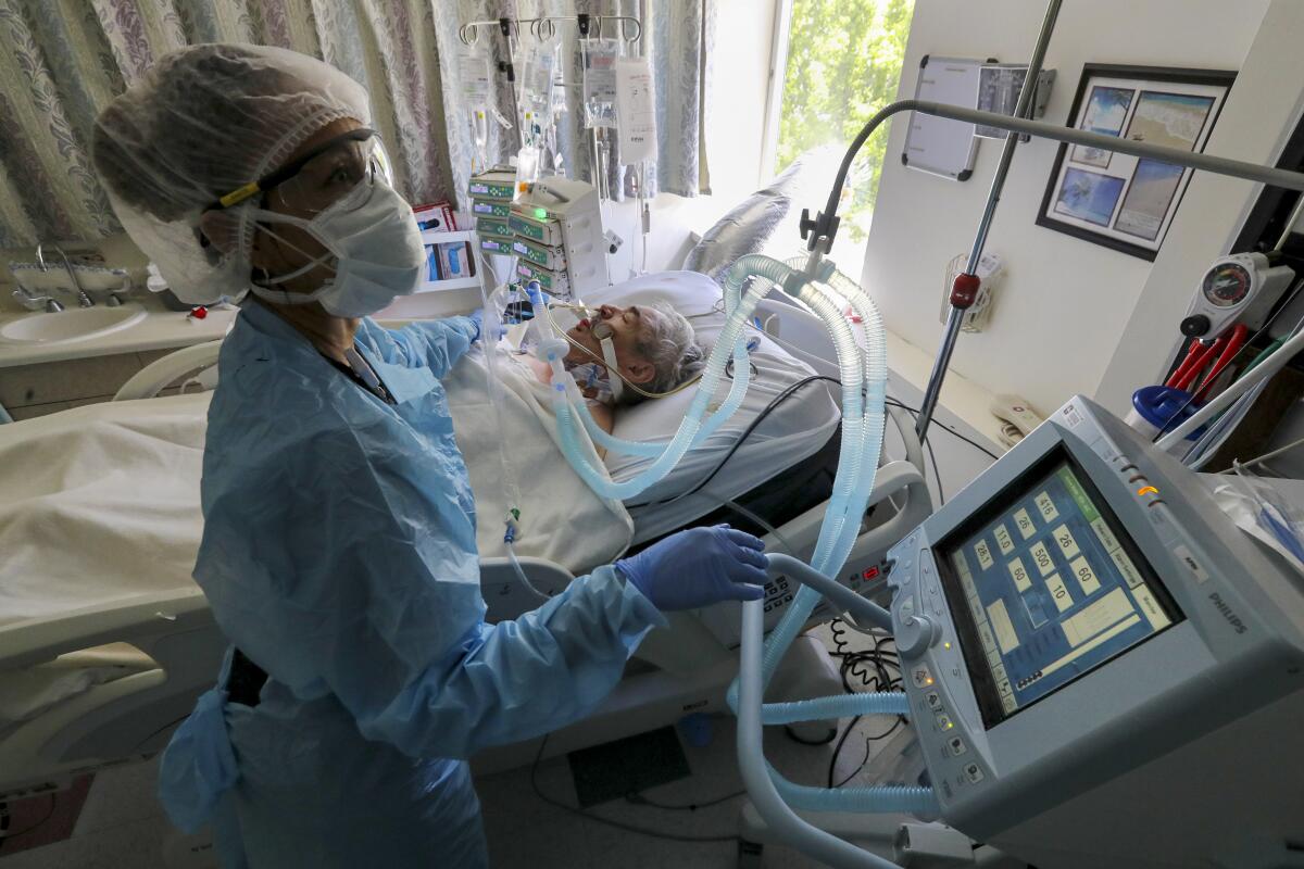 A nurse in a mask checks a machine next to an intubated patient's hospital bed