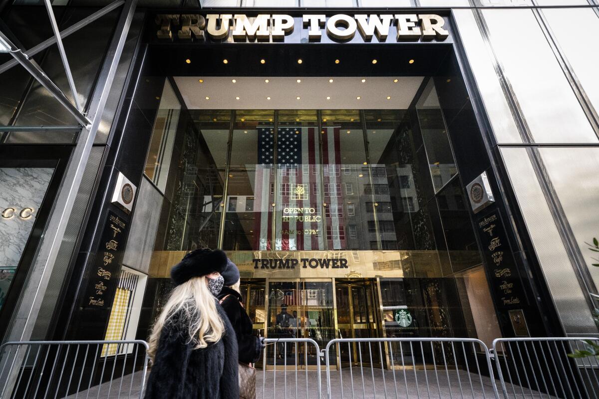 Two people walk by a towering entryway with the words "Trump Tower" and a large U.S. flag.
