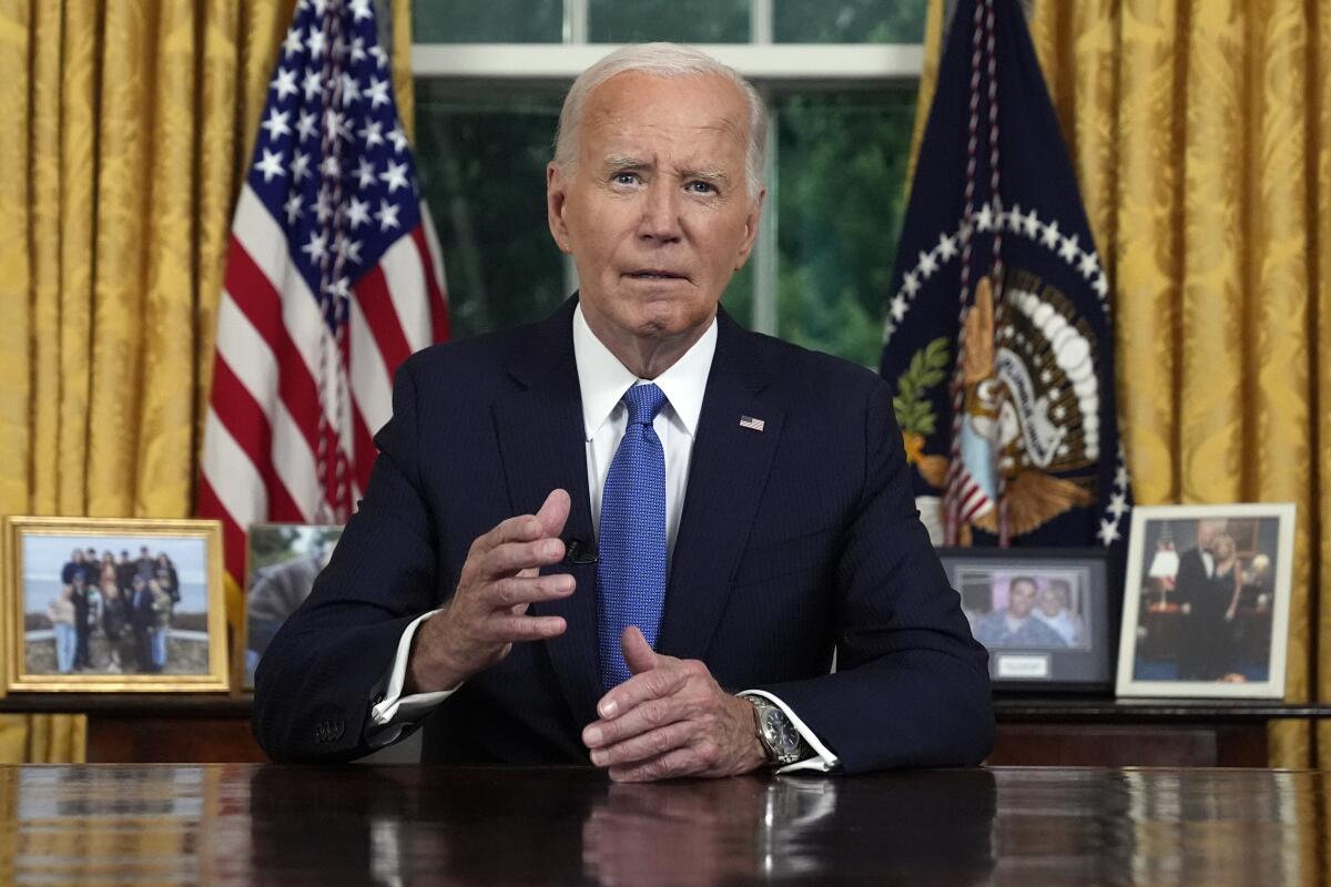 Biden gestures at a desk, with flags behind him.