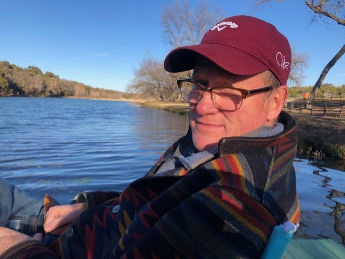 A man in a ballcap and glasses sits near a lake.