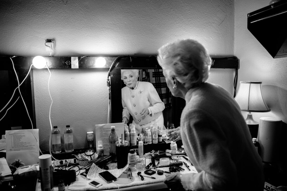 A woman in a conservative skirt suit and brooch is reflected in a dressing room mirror over a table filled with makeup.