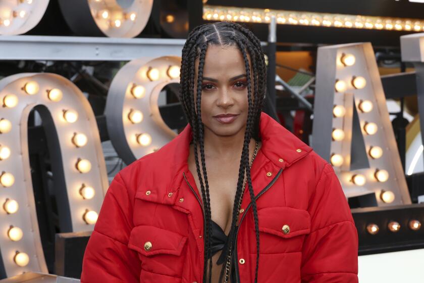 Christina Milian poses in a red coat in front of a sign made of light bulbs.