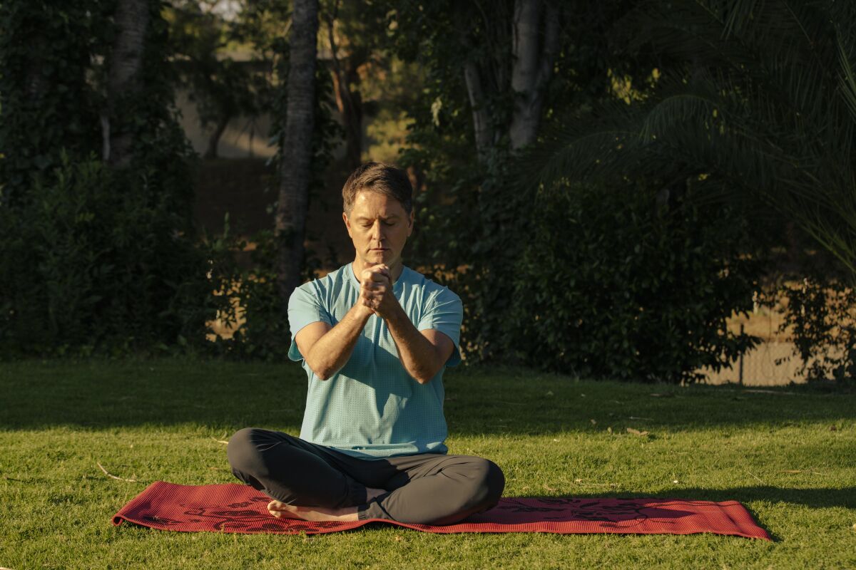 A man sits  on a yoga mat in the park, with his legs crossed and hands clasped in front of his chest.