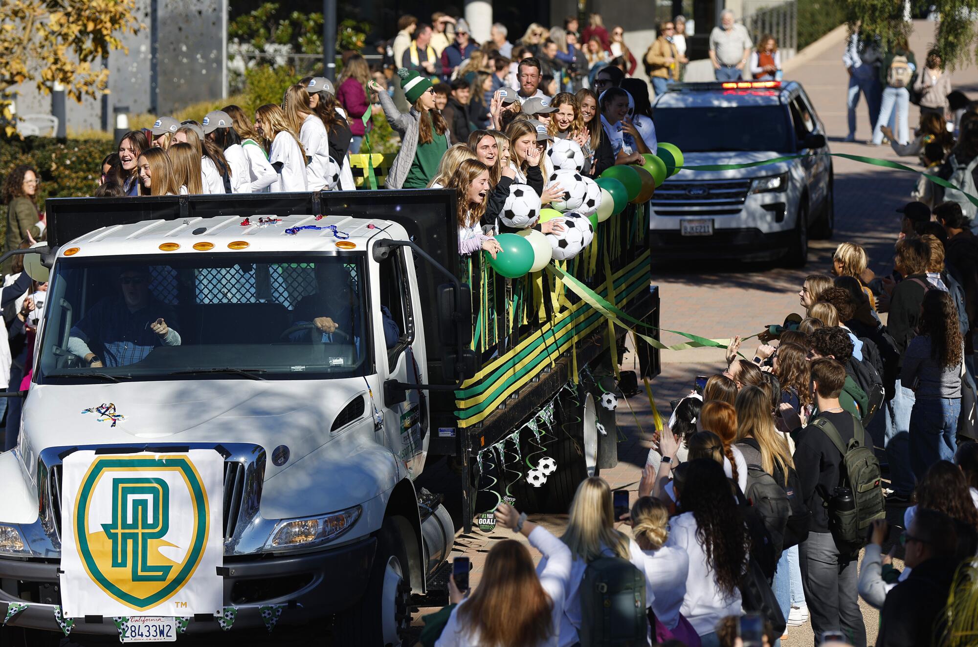 Crowds cheer as a flatbed truck full of women in soccer uniforms holding soccer balls and balloons drives down a street.