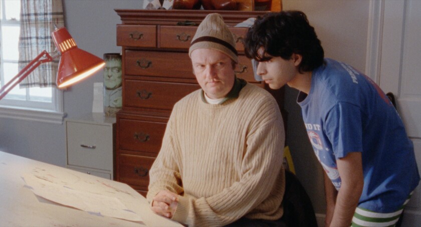 A man sits at a drafting table as a younger man looks over his shoulder at drawings taped there.