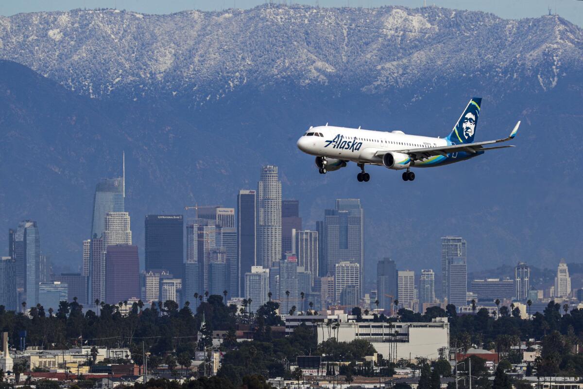 An Alaska Airlines jet flying by the downtown Los Angeles skyline against a backdrop of snow-dusted mountains