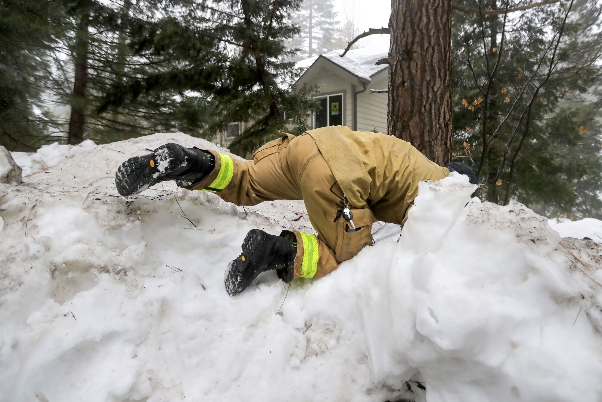 A person in a tan firefighter uniform climbs over a steep snow berm, their head disappearing behind a pile
