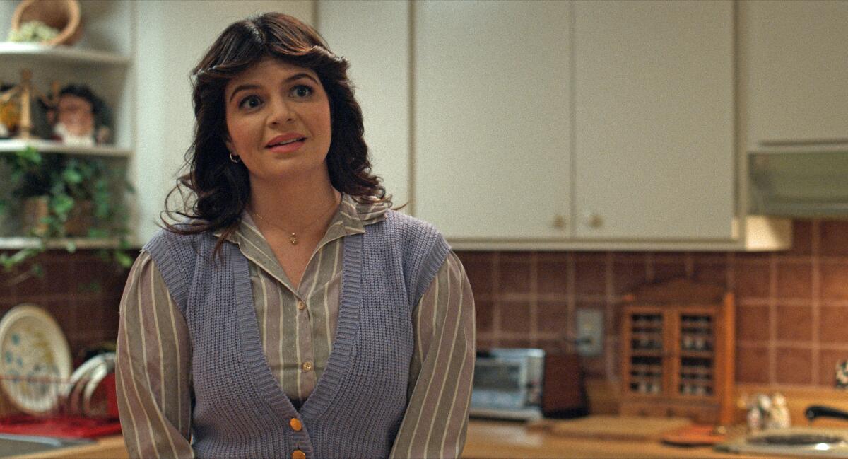 A woman in a blue sweater vest and button down stands in a kitchen